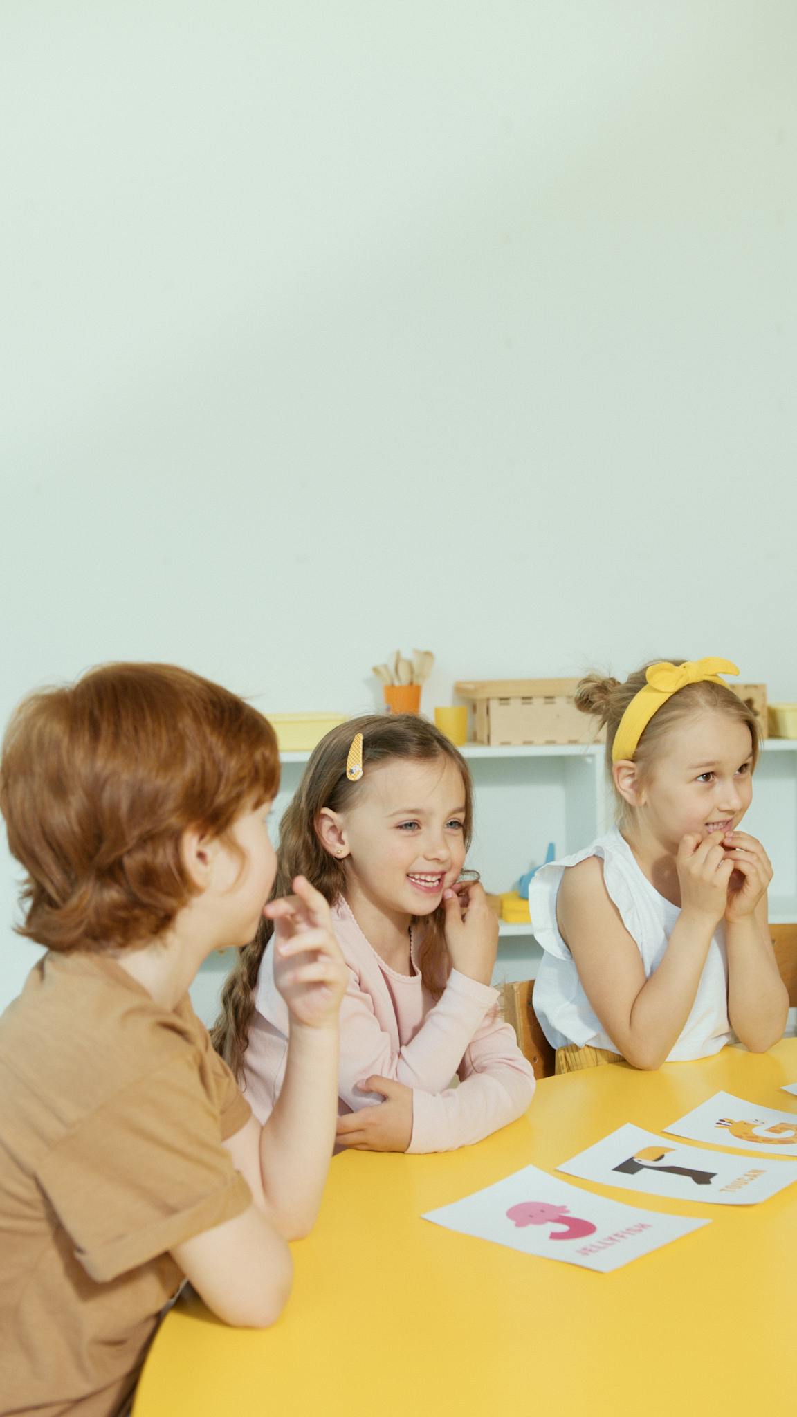 Group of children laughing and participating in a classroom setting around a yellow table.