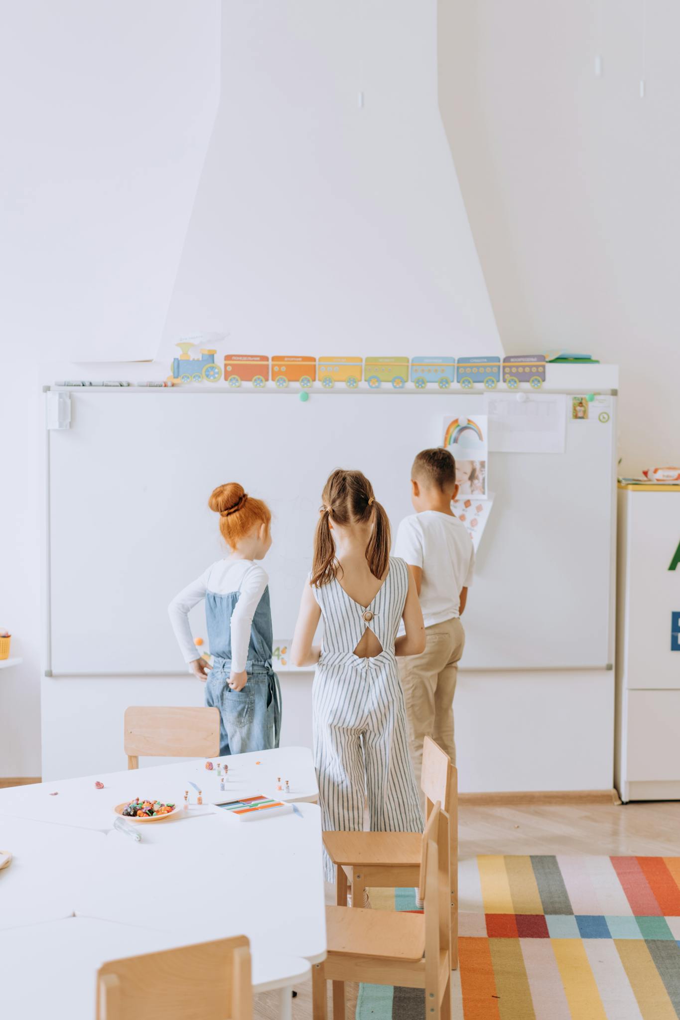 Three children in a preschool classroom engaged at a whiteboard during a learning activity.